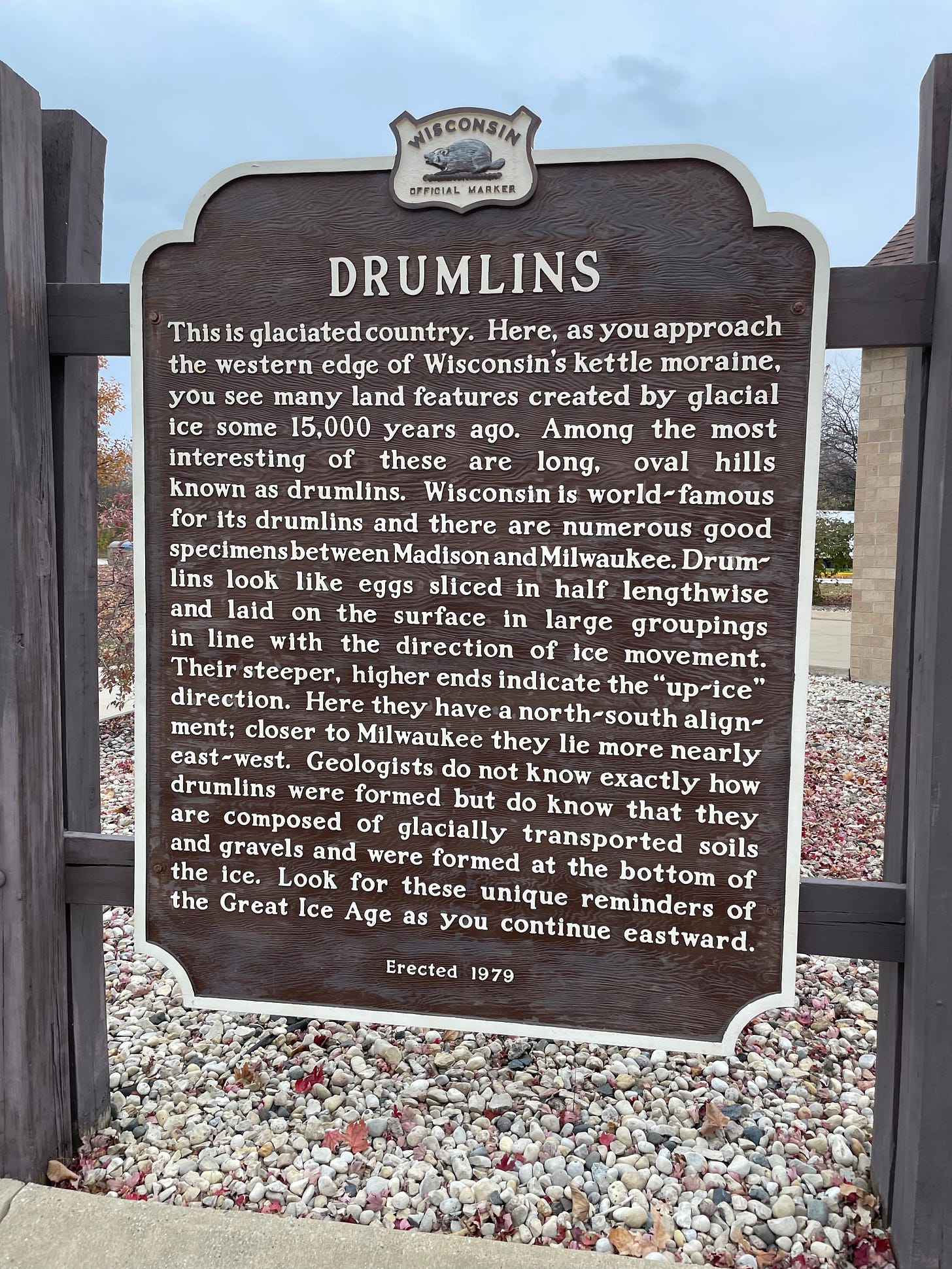 DRUMLINS: This is glaciated country. Here, as you approach the western edge of Wisconsin’s kettle moraine, you see many land features created by glacial ice some 15,000 years ago. Among the most interesting of these are long, oval hills known as drumlins. Wisconsin is world-famous for its drumlins and there are numerous good specimens between Madison and Milwaukee. Drumlins look like eggs sliced in half lengthwise and laid on the surface in large groupings in line with the direction of ice movement. Their steeper, higher ends indicate the “up-ice” direction. Here they have a north-south alignment; closer to Milwaukee they lie more nearly east-west. Geologists do not know exactly how drumlins were formed but do know that they are composed of glacially transported soils and gravels and were formed at the bottom of the ice. Look for these unique reminders of the Great Ice Age as you continue eastward. (Erected 1979)