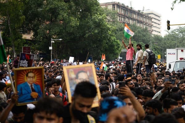 A large crowd of people fills a street. Many hold framed portraits and Indian flags; one person stands elevated, holding a large flag.