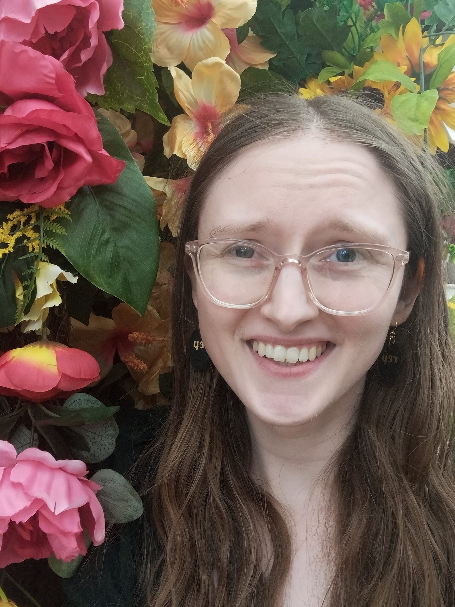 A selfie of Kandi Zeller (she/her), a white woman with long brown hair and pink glasses, with a backdrop of silk flowers that are hanging up in a craft store aisle