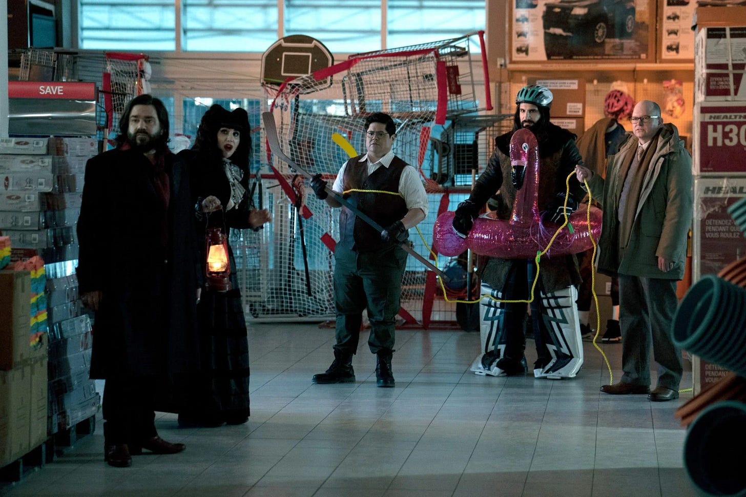 Five oddly dressed people stand frozen looking off to the left looking surprised and confused while standing amongst boxes and sporting goods in a big warehouse space.