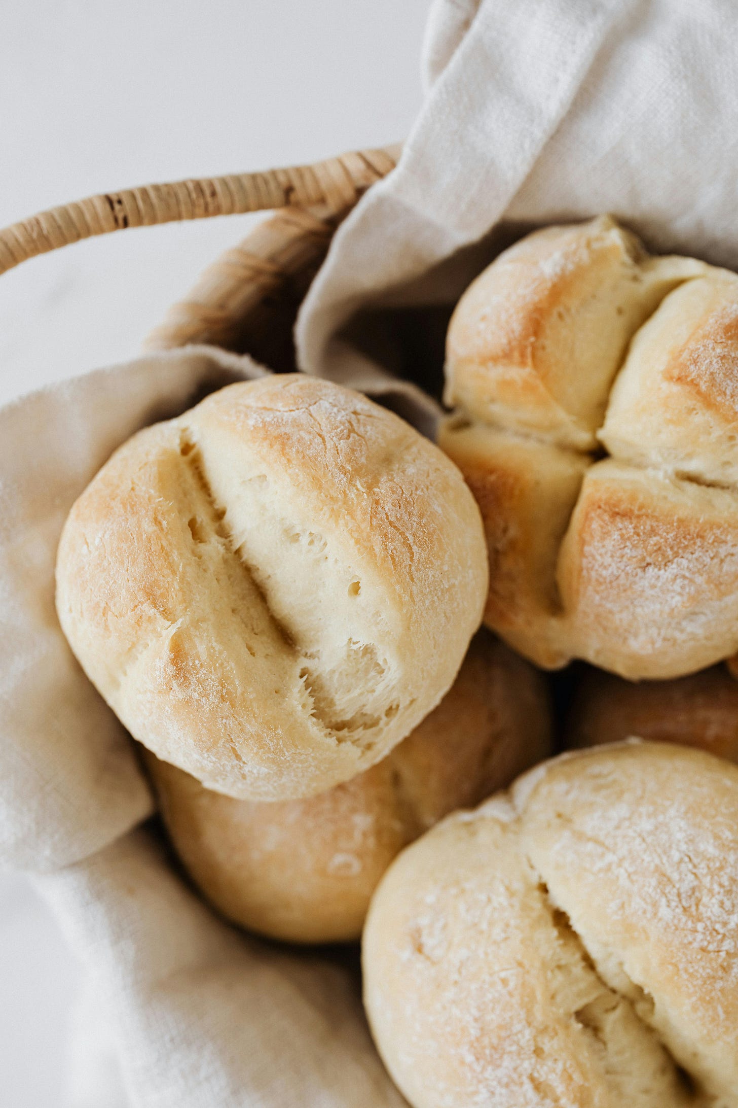 white bread buns with a crispy looking exterior laying in a wicker basket we can just see the handle of on a white background. buns have a mark down the middle looking like they were split in half before baking and are on top of a white cloth in the basket.
