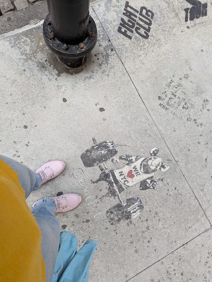 1. two black garbage bins filled with pansie flowers in soil are sitting on a sidewalk. In the background there is a fire hydrant. 2. POV of legs in urban sneakers and jeans with a stencil graffiti of a weightlifting rat wearing a "We LOVE NYC" t-shirt.