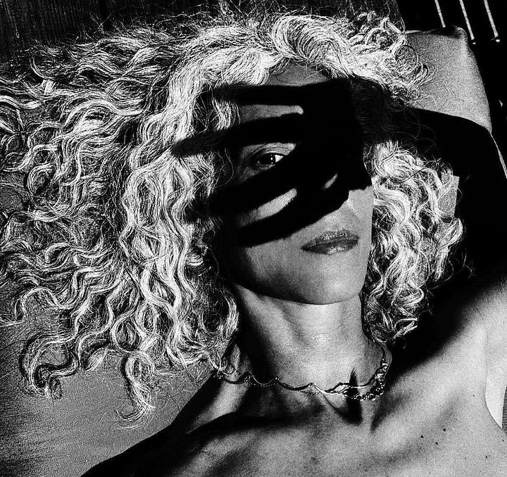 Black-and-white self-portrait of a woman holding two large flower heads in front of her eyes like a mask. The flowers obscure her face, leaving only her lips and the strong lines of her neck and shoulders visible. The image is intimate, surreal, and striking in its simplicity. Black-and-white self-portrait of a woman lying down with her curly hair fanned out, partially covered by deep shadows cast from her own hand. The image plays with light and shadow to create a sense of mystery and emotion, capturing both strength and softness.