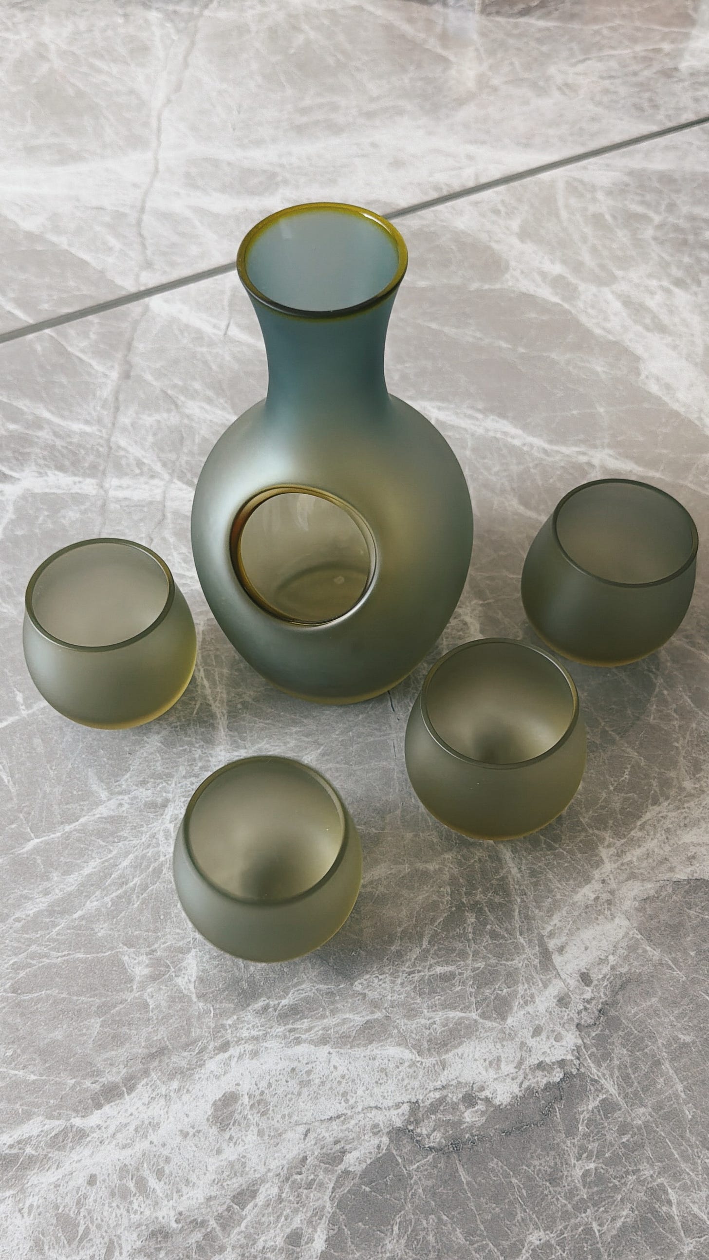 A frosted green glass sake carafe and four curved cups arranged on a gray veined stone surface A frosted green glass sake carafe and four curved cups arranged on a gray veined stone surface
