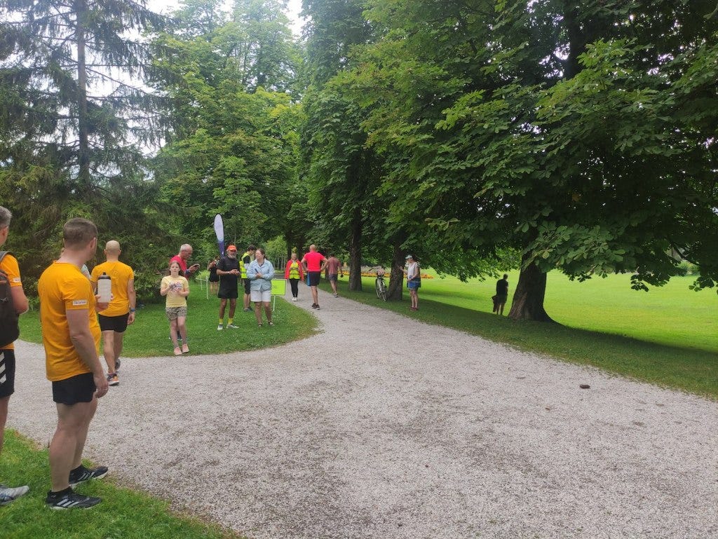 People gathered at the finish line, on the grass where several gravelled paths meet. Trees and grass are on each side.