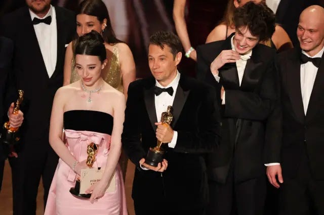 Equipe de 'Anora' no palco, incluindo atriz e diretor do filme segurando estatuetas