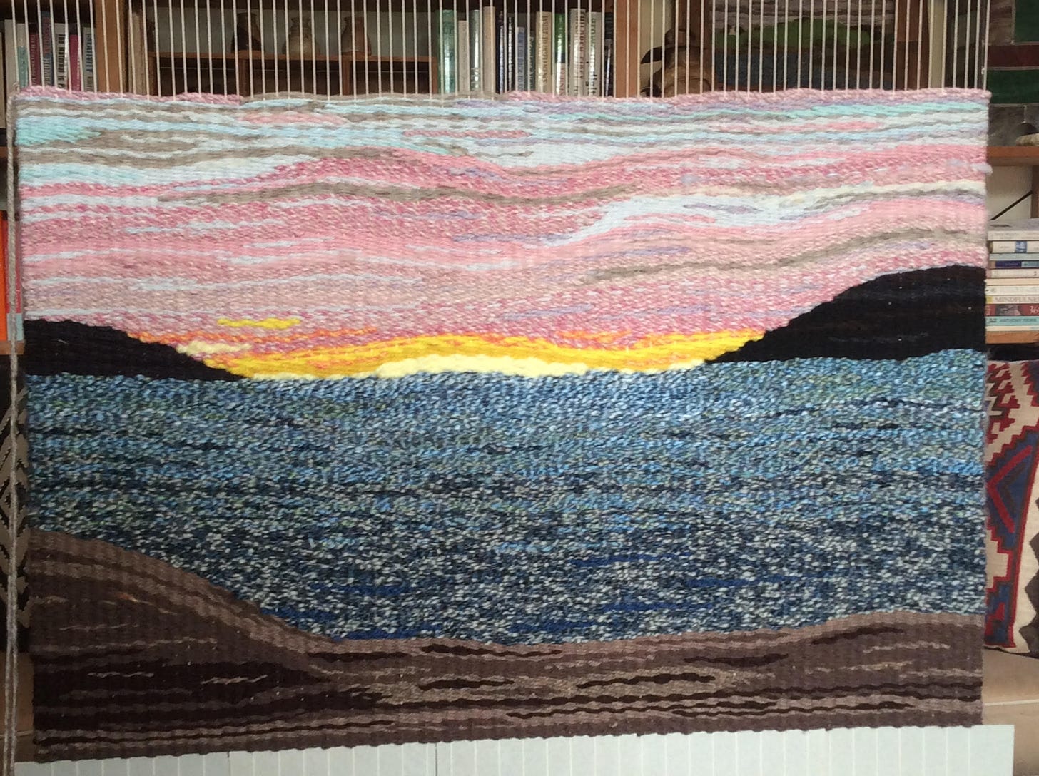 A woven tapestry ready to be cut off the loom. It includes a beach in shades of brown, an ocean in shades of blue, cliffs in the background, a setting sun with pink clouds above, clearing to blue at the top.