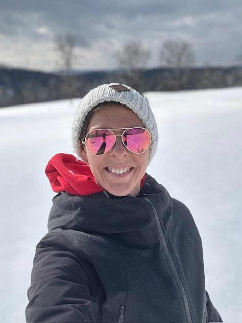 Woman makes a snow angel. Woman in a beanie cap, black jacket, and pink aviator glasses smiles in front of snow. A giant snow person hi-fives a lesbian with it's twig arm.