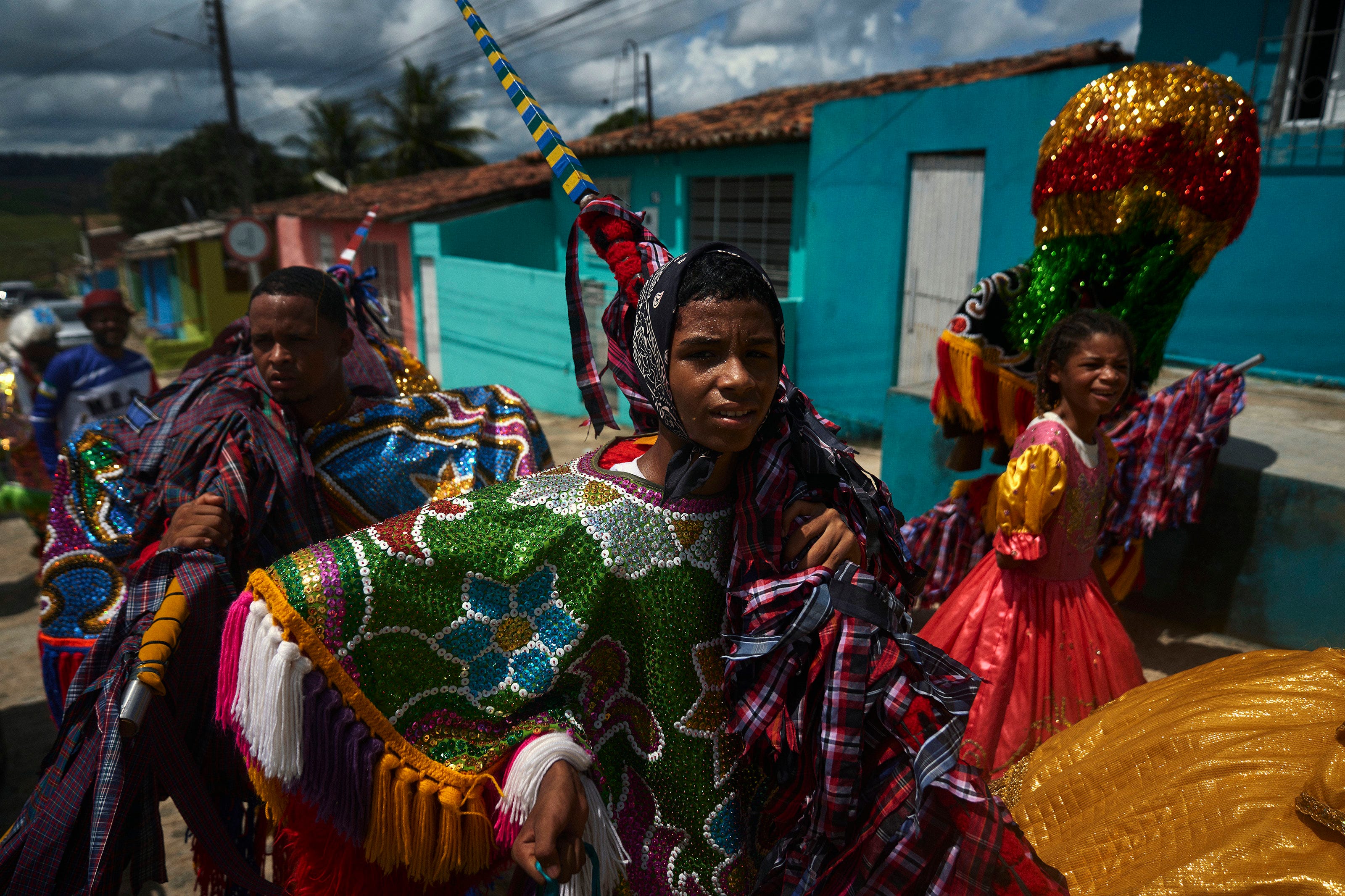 Three people in colorful, sequined costumes participate in a vibrant street parade; bright blue and turquoise buildings are seen in the background under a partly cloudy sky.