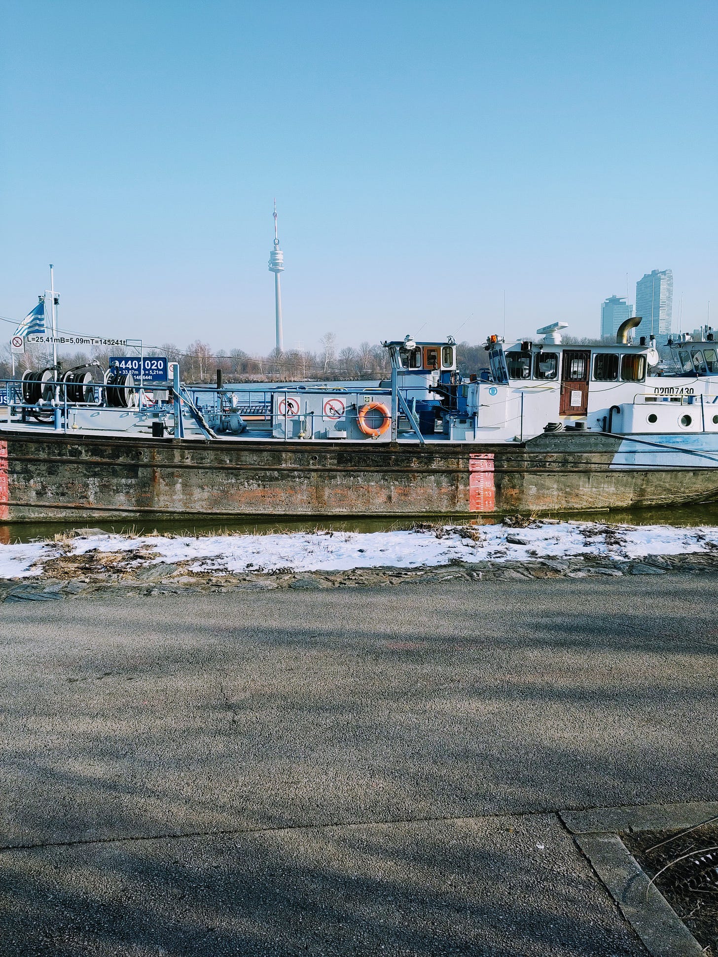 The picture was taken outside on a cold winter morning. The skies are blue and there is a boat anchoring in the front, on the Danube. In the back, the Donauturm in Vienna is visible.