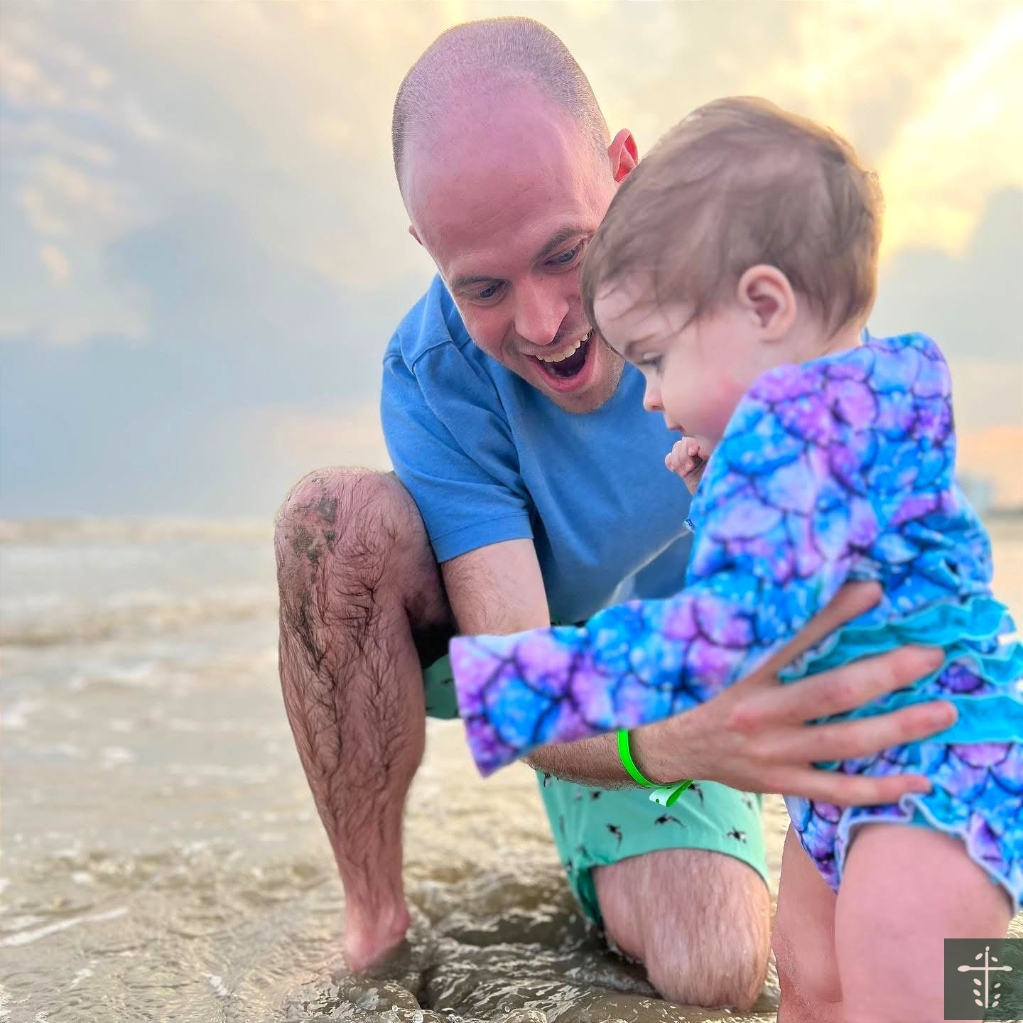 A father kneeling in the surf, smiling at his young daughter as she wades into the water.
