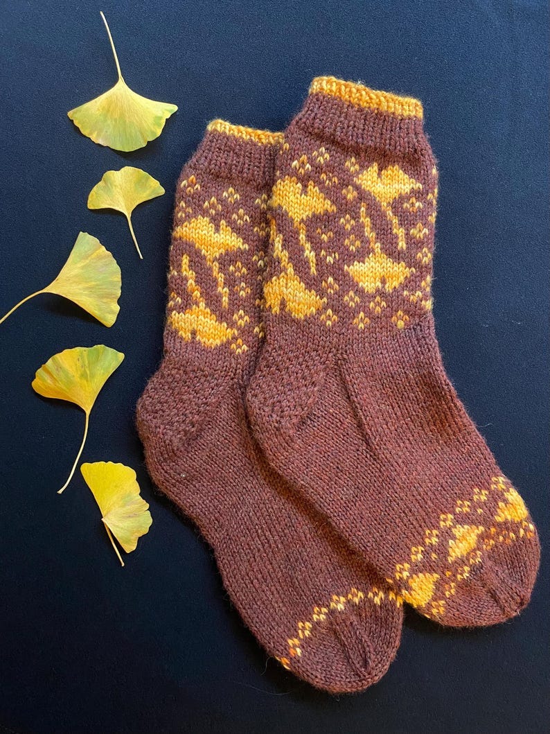 May include: Pair of hand-knitted socks in a deep brown color with a Fair Isle pattern. The pattern features yellow-gold ginkgo leaves. The socks are shown against a dark blue background with several yellow-gold ginkgo leaves.