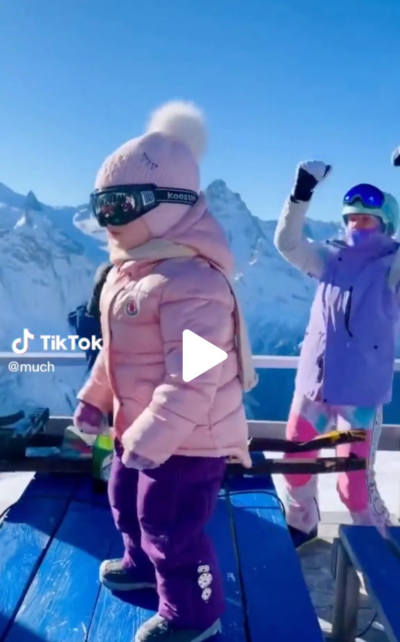 A little girl in a ski outfit on top of a picnic table with an adult in the background with her hands in the air. A little girl in a ski outfit on top of a picnic table with an adult in the background with her hands in the air.