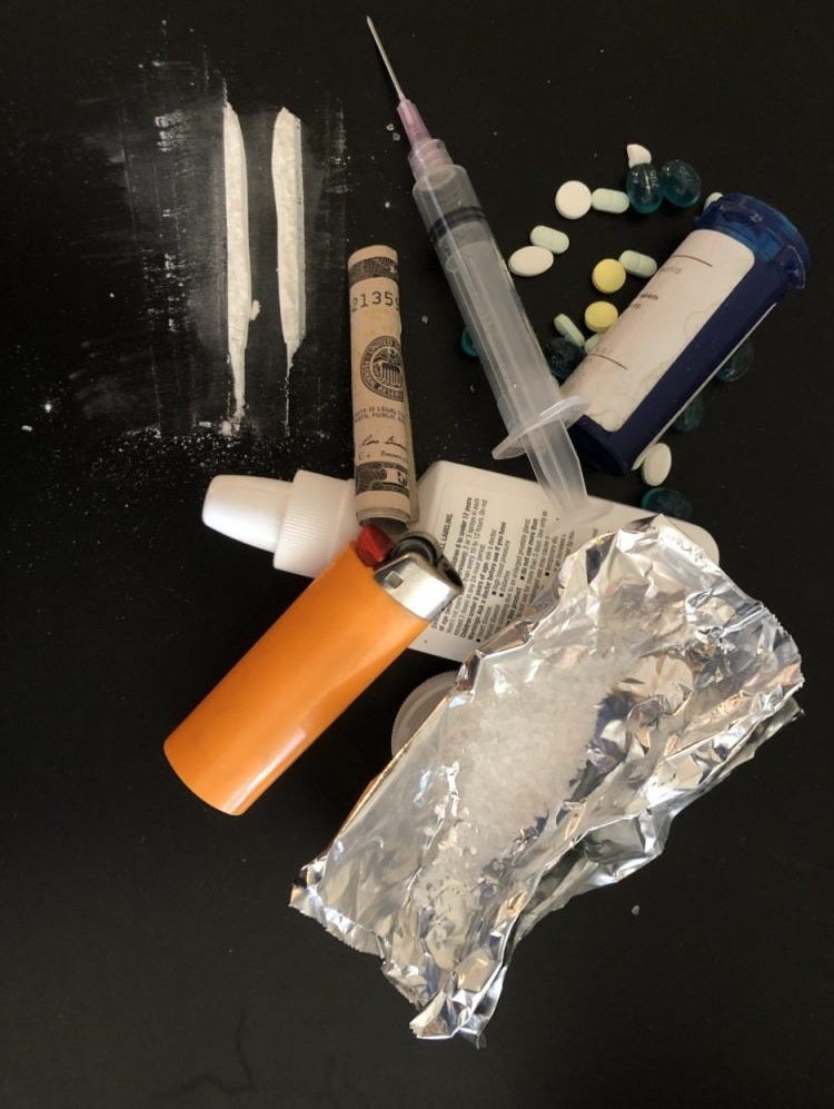 An assortment of pills, syringes with needs, lines of powdered drug, a lighter, a rolled dollar bill, and crystals on tin foil. 