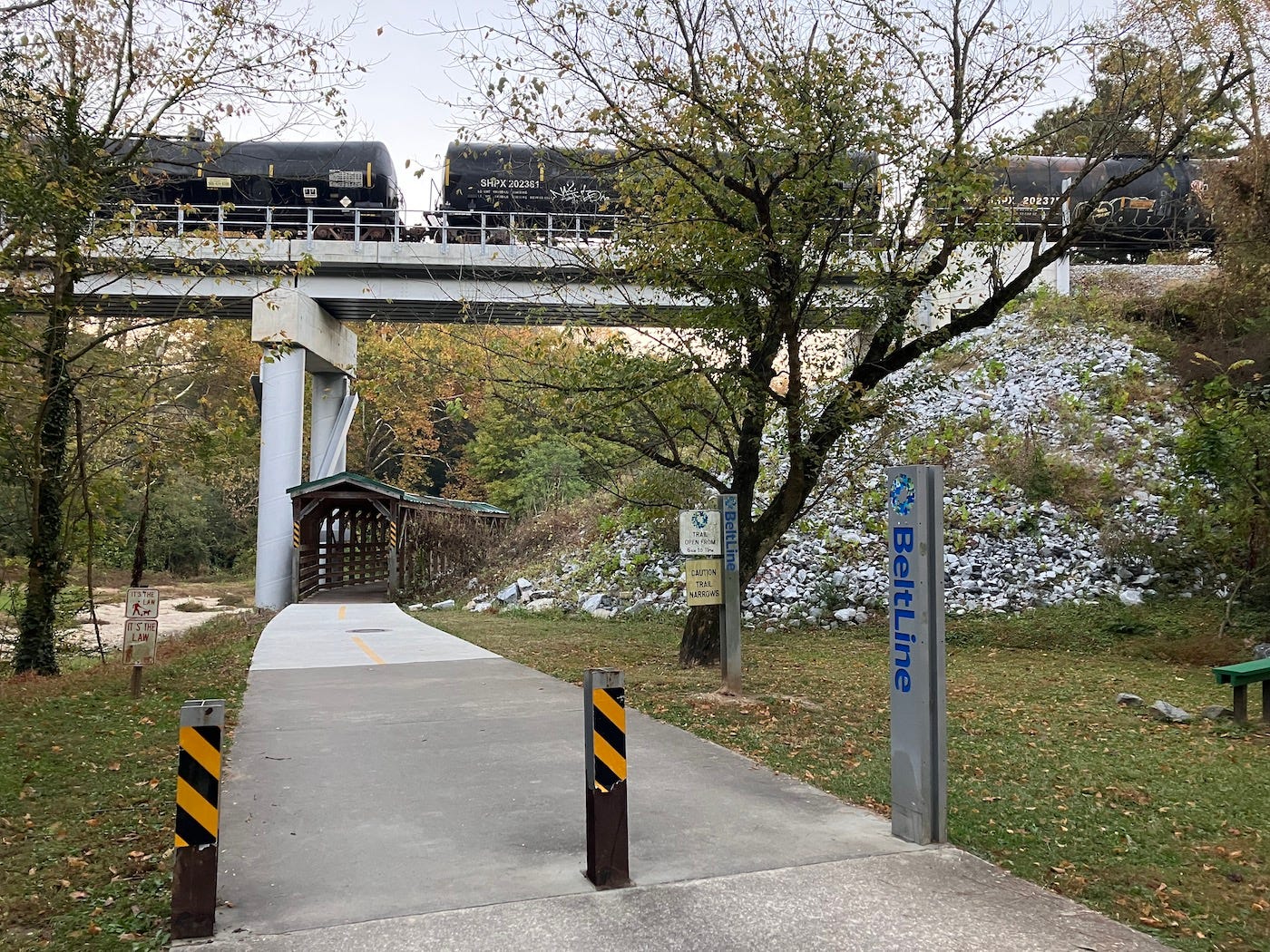 Concrete trail passing through a wooden covered bridge for pedestrians, with a freight train on the bridge above