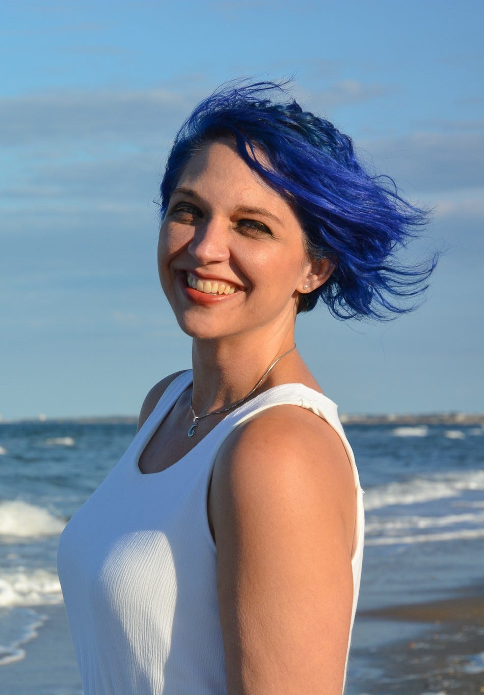 A woman with blue hair stands on the beach and smiles at the camera