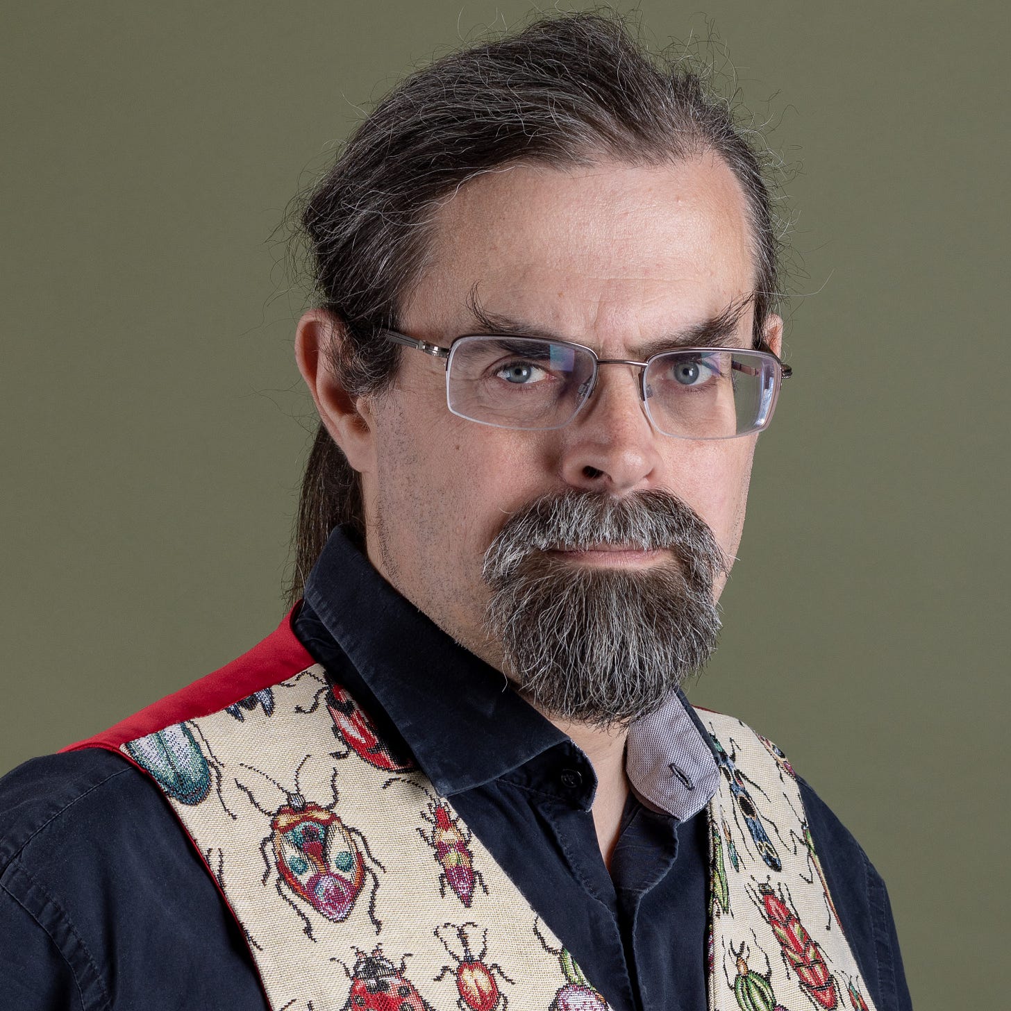 Studio portrait of a man with glasses, tied-back dark hair, and a grey beard, wearing a black shirt and a cream waistcoat patterned with colourful beetles.
