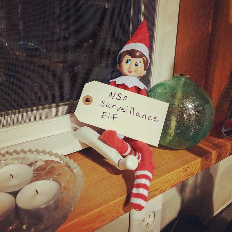 A photo of an Elf on the Shelf toy doll sitting on a windowsill and holding a small sign, which reads "NSA Surveillance Elf."
