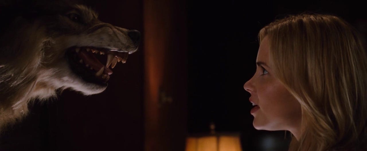 A blonde woman's side profile is staring directly at a taxidermy wolf's side profile. The wolf is snarling, teeth bared.