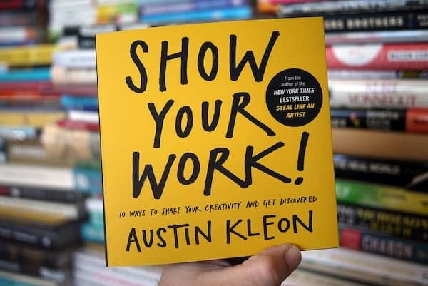 A person holding the book 'Show Your Work!' by Austin Kleon, with a backdrop of colorful bookshelves. A person holding the book 'Show Your Work!' by Austin Kleon, with a backdrop of colorful bookshelves.