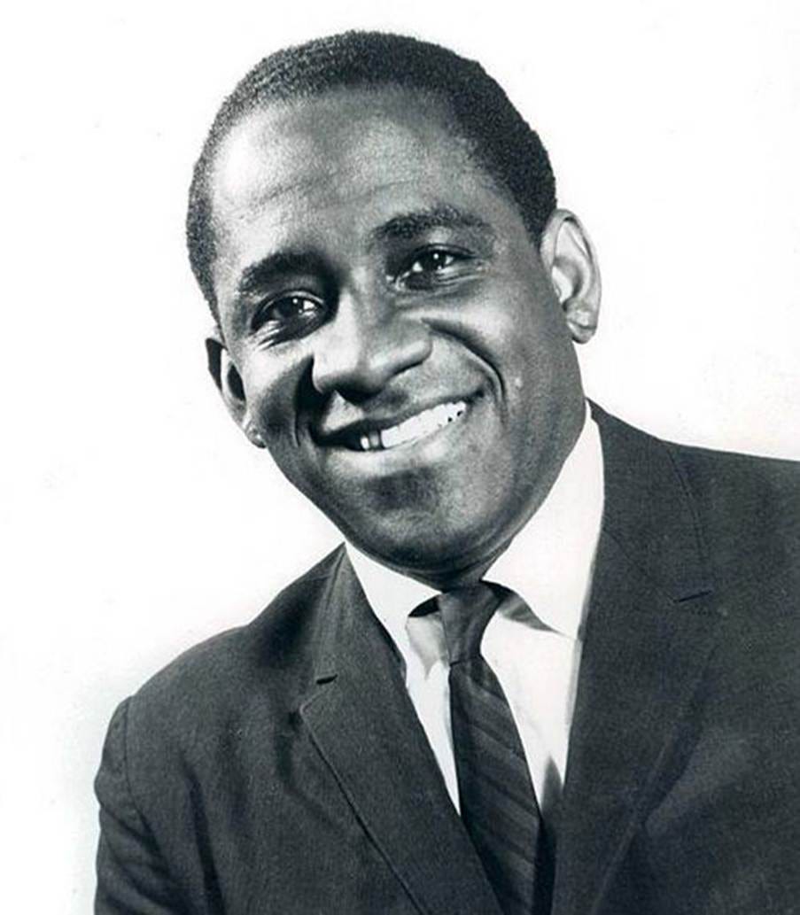 Black and white photo of a smiling Mongo Santamaría wearing a suit, white shirt, and tie, with short hair. Picture Credit: Wikipedia