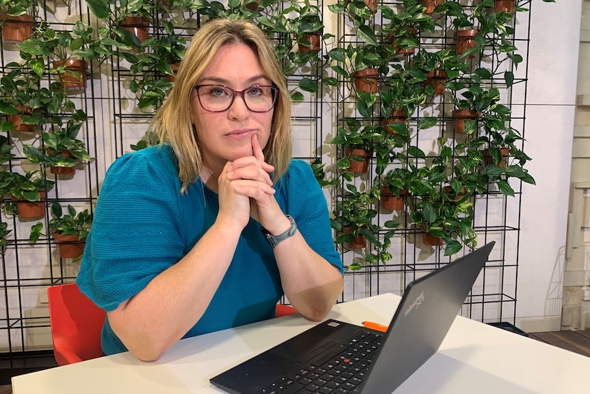 A woman in a blue shirt and glasses sits at a desk with a laptop A woman in a blue shirt and glasses sits at a desk with a laptop