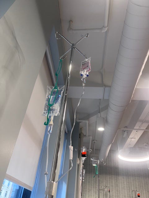Shot of clear IV bags hanging on a metal hook. In the background, you can see more hooks with an IV bag of dark liquid.