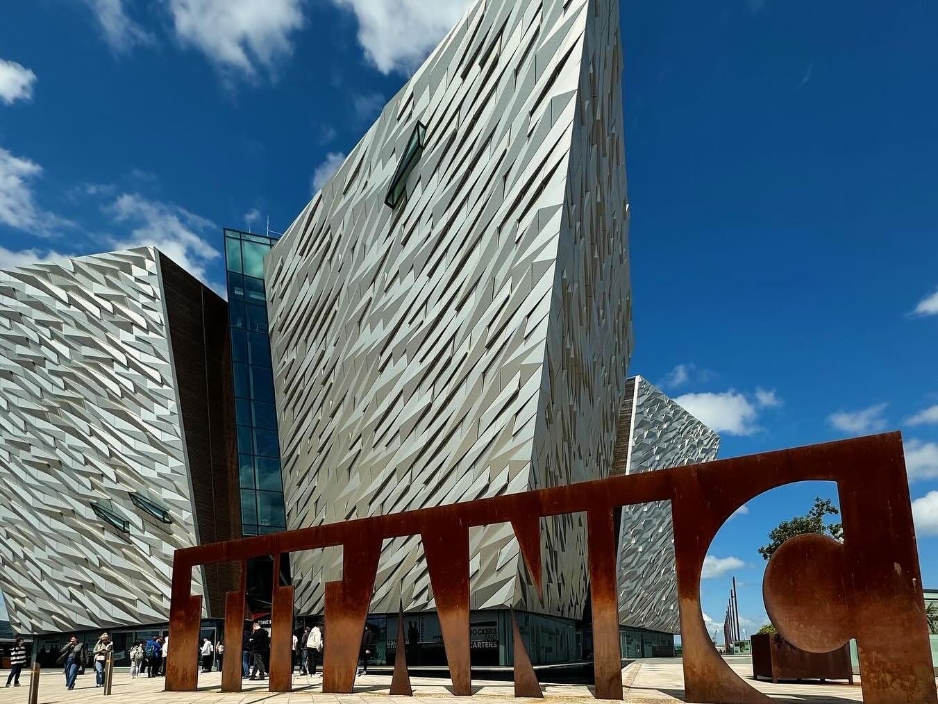 Modern architectural structure with angular, textured facades, featuring a large "TITANIC" sign in the foreground. Visitors gather outside under a blue sky.