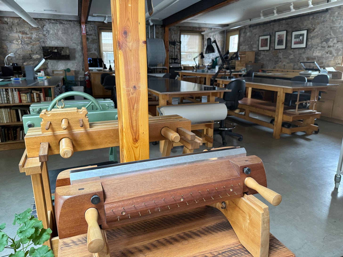 A big room with stone brick walls and concrete floors. In the room are lots of wooden tables and contraptions used for book binding classes.