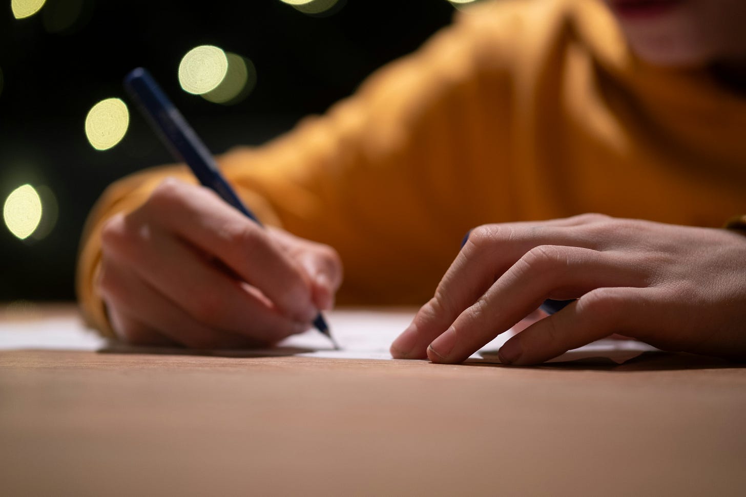 Close-up photograph of hands writing with a pen on paper, with warm golden bokeh lights creating a soft, luminous background. Close-up photograph of hands writing with a pen on paper, with warm golden bokeh lights creating a soft, luminous background.