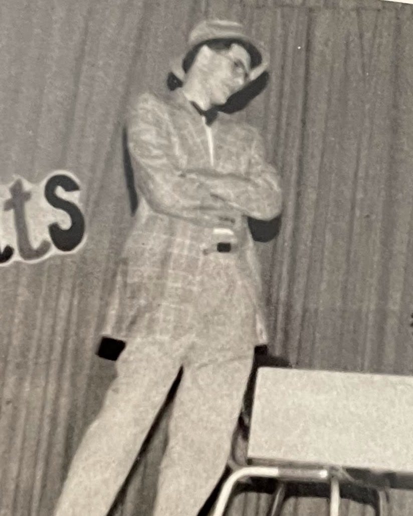 A black-and-white photograph of a young person on stage, wearing a plaid suit, bow tie, and fedora-style hat. They stand with arms crossed, leaning slightly to one side, in front of a curtain with partially visible lettering. A metal folding chair sits nearby.