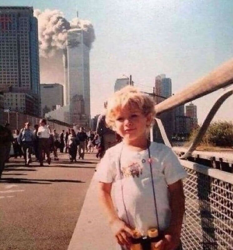 A young child stands smiling in the foreground with binoculars around their neck. In the background, smoke billows from the World Trade Center towers in New York City, as people walk and look toward the scene.