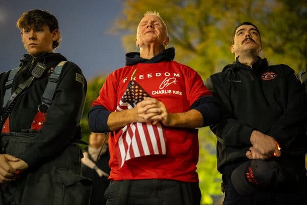 Three men stand with their hands clasped in prayer. One of them, Joe Schoendorf, is holding an American flag and wearing a red T-shirt with the words, “Freedom Charlie Kirk.”