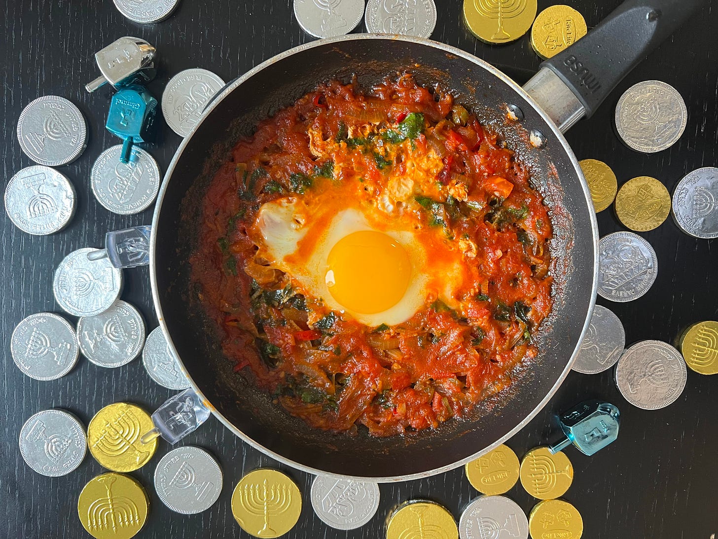 Shakshuka with a sunny-side-up egg in the center, cooked in a rich tomato sauce with spinach and spices. The pan is placed on a black surface, surrounded by silver and gold Hanukkah gelt coins and blue dreidels. Part of Liat Portal’s Foodie Disorder journey on The Liat Show.