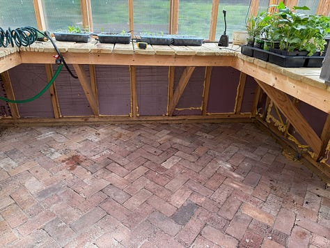 Alt text: Three side-by-side photos of a greenhouse interior during construction. The images show newly built wooden raised planting beds arranged around the perimeter of the space, with a herringbone-pattern brick floor installed throughout the center walkway. The greenhouse framing and clear wall panels are visible, and the beds are unfinished, showing the structural supports beneath the bench tops. The photos capture different angles of the same space, highlighting the layout of the raised beds and brick flooring.