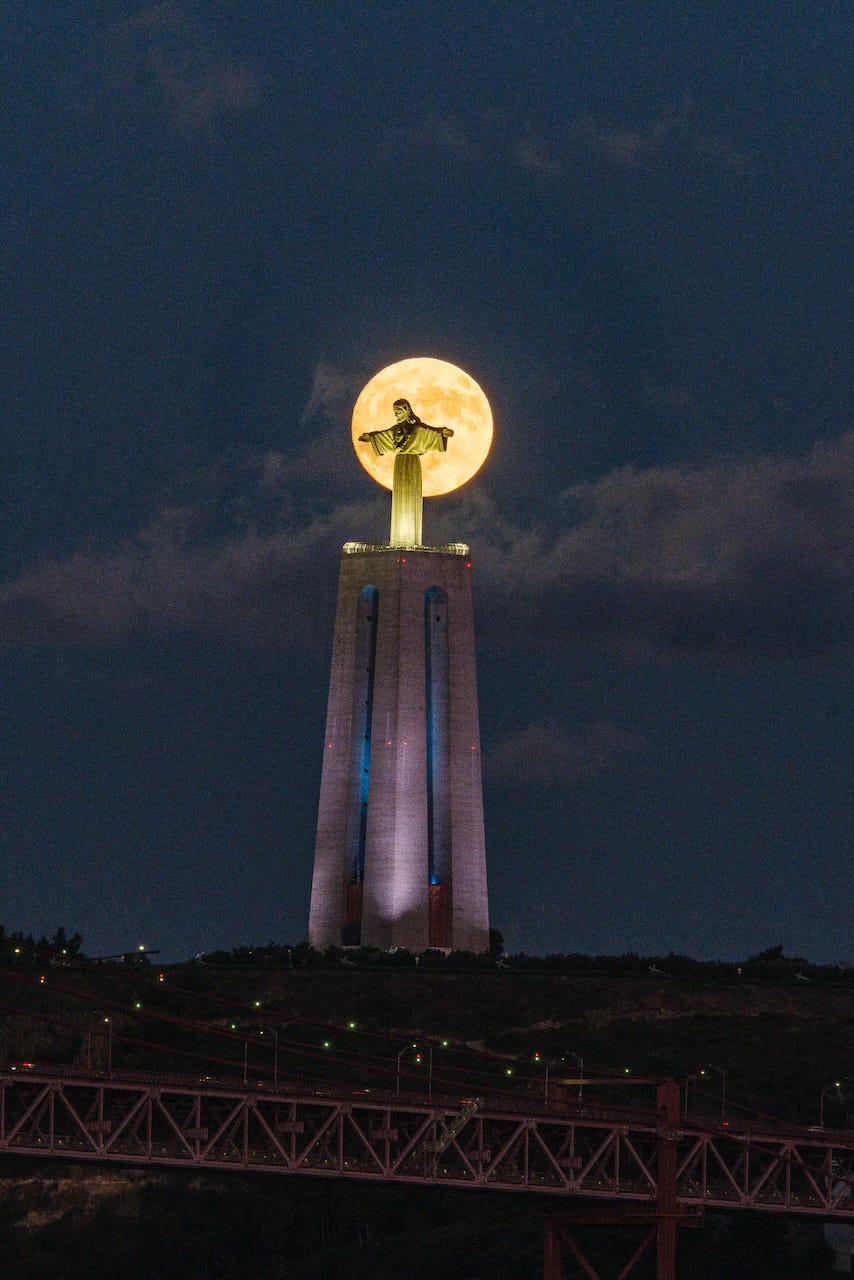 The full moon beyond the statue of Cristo Rei in Lisbon, Portugal