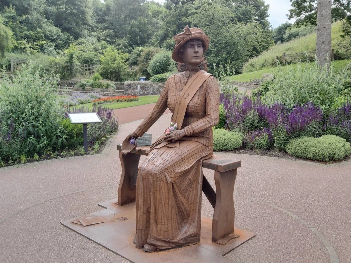 Wooden sculpture of a seated woman in a hat, surrounded by a lush garden with flowers and greenery.