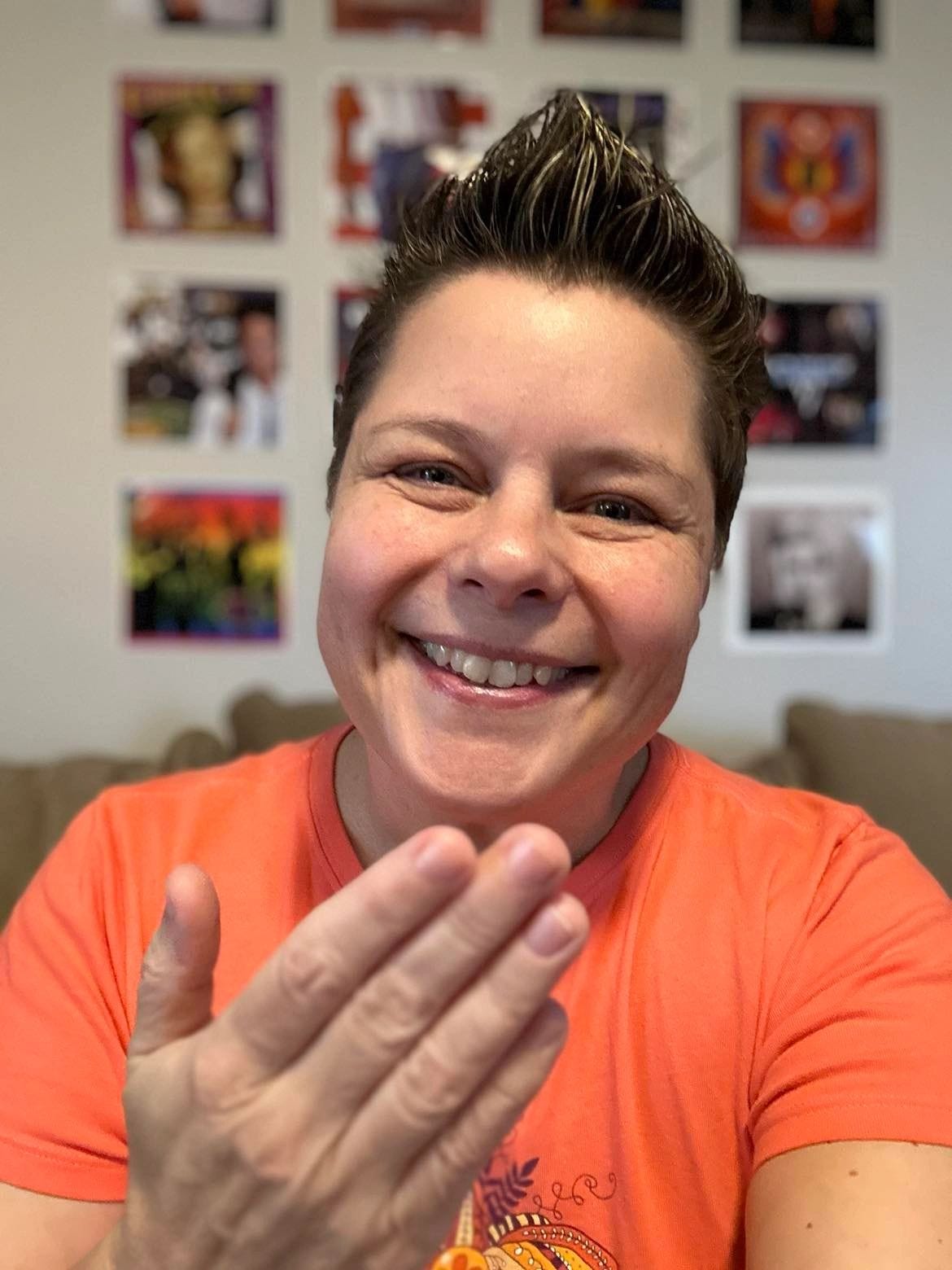 Chris Davis signs thank you in ASL. She shows a friendly smile and wears an orange shirt. Her signature '80s album wall is in the background.