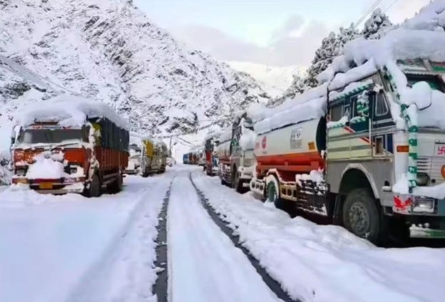 Line of colorful trucks stuck on a snow-covered mountain road surrounded by steep white peaks under a cloudy sky.