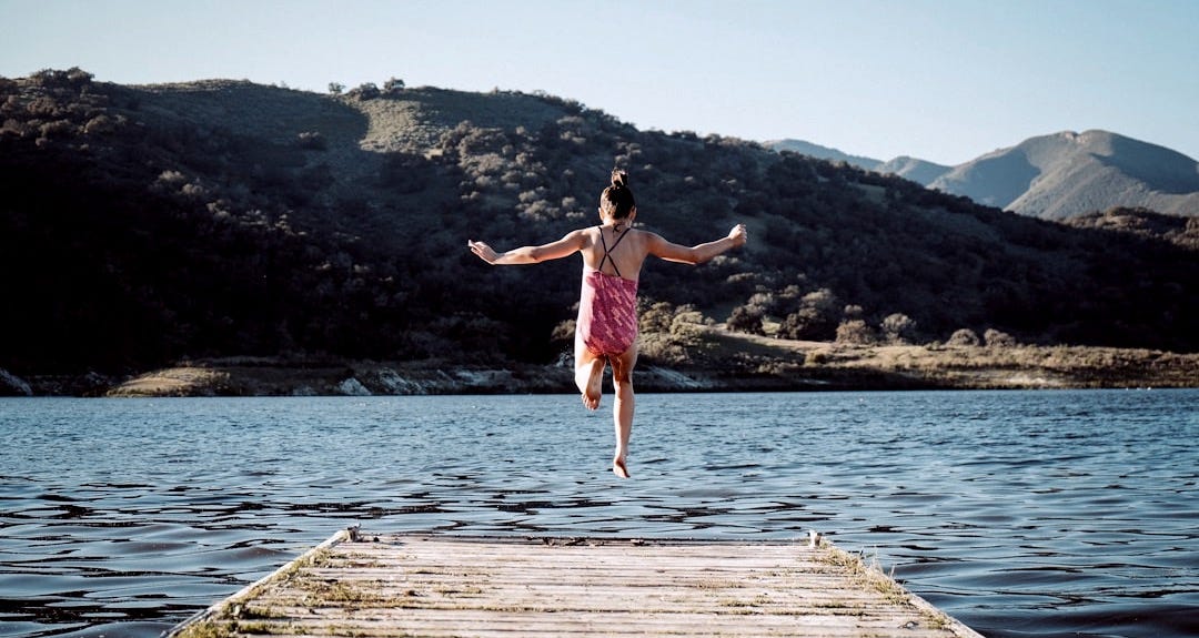 woman wearing pink top jumping towards water during daytime