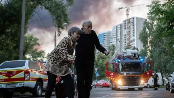 An elderly couple appear to be walking towards shelter in Kyiv. A fire engine, tall buildings and smoke are seen in the background.