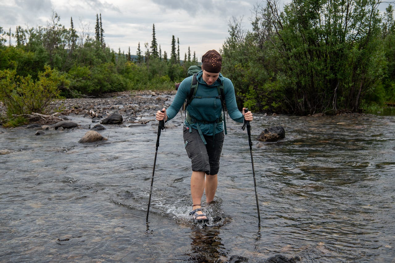 A hiker in a teal top and rolled shorts wades a shallow, clear creek in sandals, leaning on two trekking poles, a pack on her back and her head down watching her footing. Spruce and green willow line both banks under a soft grey sky.