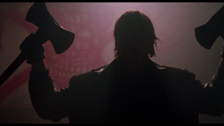 Brian Thompson as the Night Slasher in Cobra 4K UHD – Silhouette of the Night Slasher raising two axes, backlit by red light and skull graffiti in the Arrow Video restoration.