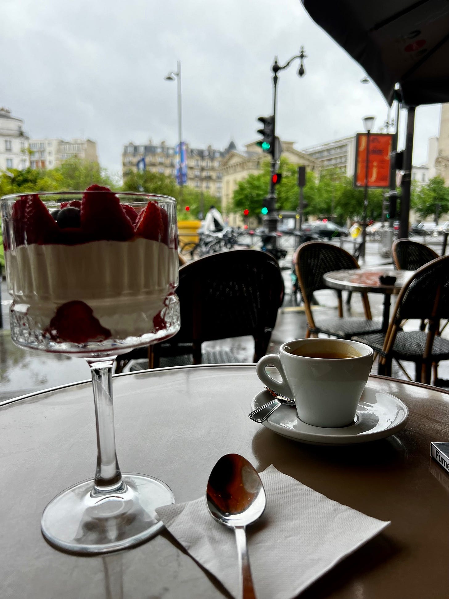 A glass of layered dessert with berries and cream sits next to a white coffee cup on a café table overlooking a busy Parisian street with traditional Haussmann-style buildings. A glass of layered dessert with berries and cream sits next to a white coffee cup on a café table overlooking a busy Parisian street with traditional Haussmann-style buildings.