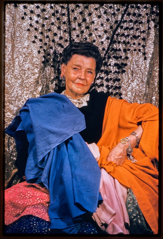 Portrait of Barbara Karinska in front of glitter curtain with blue, orange, and pink fabric draped over her. Her left hand is positioned on her hip. Portrait of Barbara Karinska in front of glitter curtain with blue, orange, and pink fabric draped over her. Her left hand is positioned on her hip.