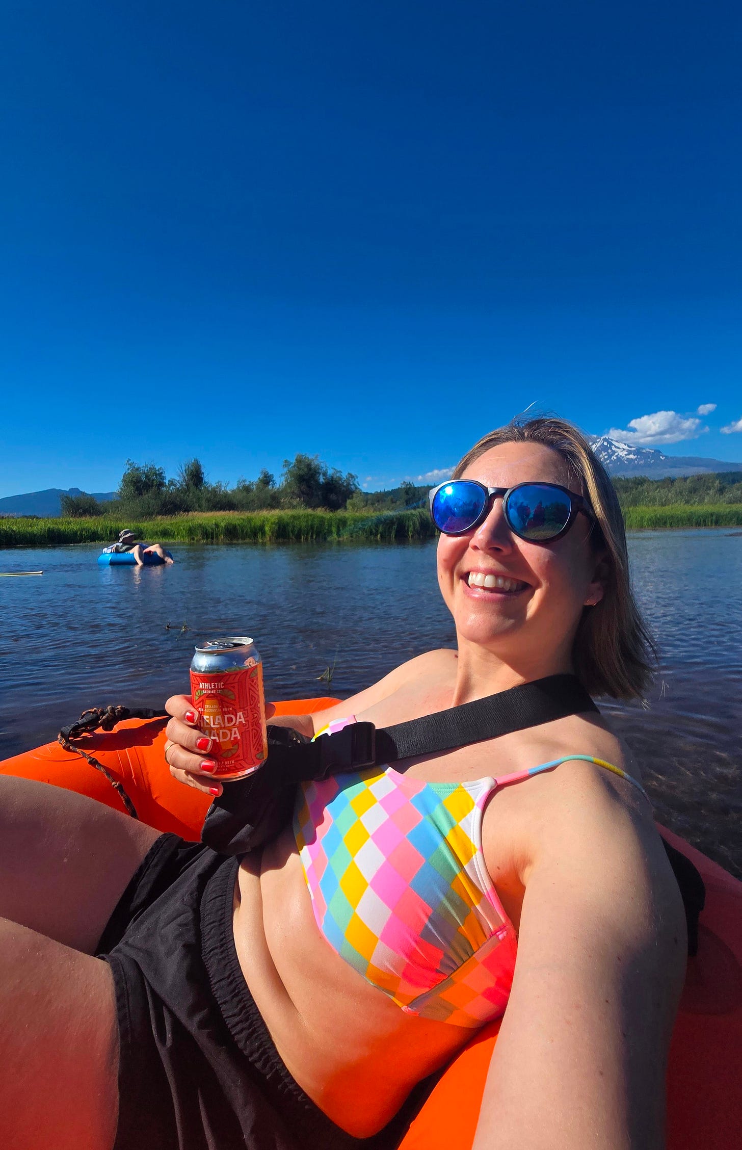 Cassidy sitting in an orange innertube floating on a creek, with blue sky and a mountain behind her, and a non-alcoholic beer in her hand