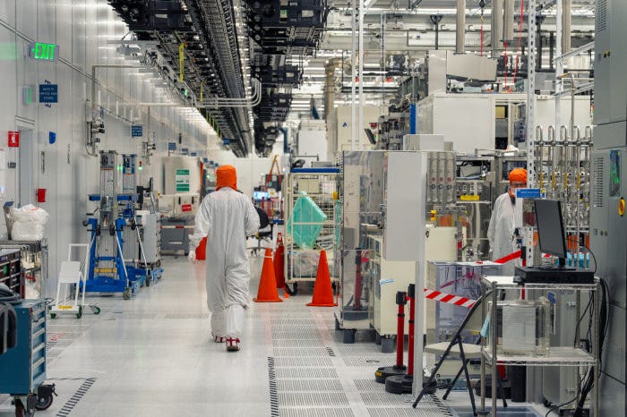 A person in a cleanroom suit and orange hood walks through an R&D fabrication facility.