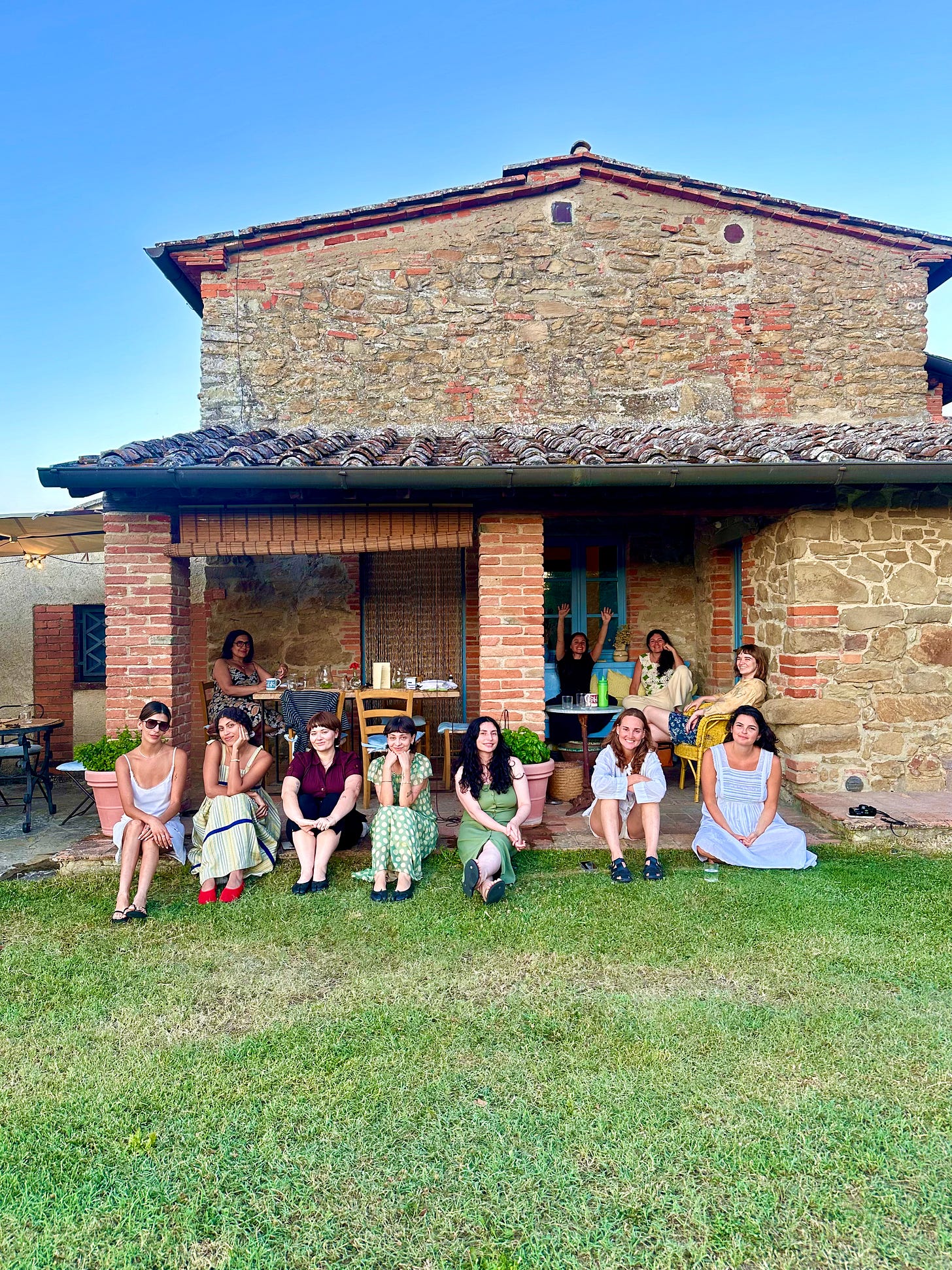A group of people sitting and relaxing on grass in front of a rustic stone house with brick columns and a tiled roof. Some sit on the lawn while others are on the porch, smiling casually in summer clothing under a clear blue sky