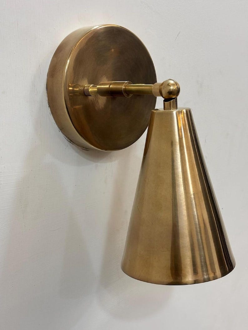 May include: A brass wall sconce with a circular backplate and a conical shade. The sconce has a warm, golden hue and is mounted on a white wall. The shade is cone-shaped and angled downward. May include: A brass wall sconce with a circular backplate and a conical shade. The sconce has a warm, golden hue and is mounted on a white wall. The shade is cone-shaped and angled downward.