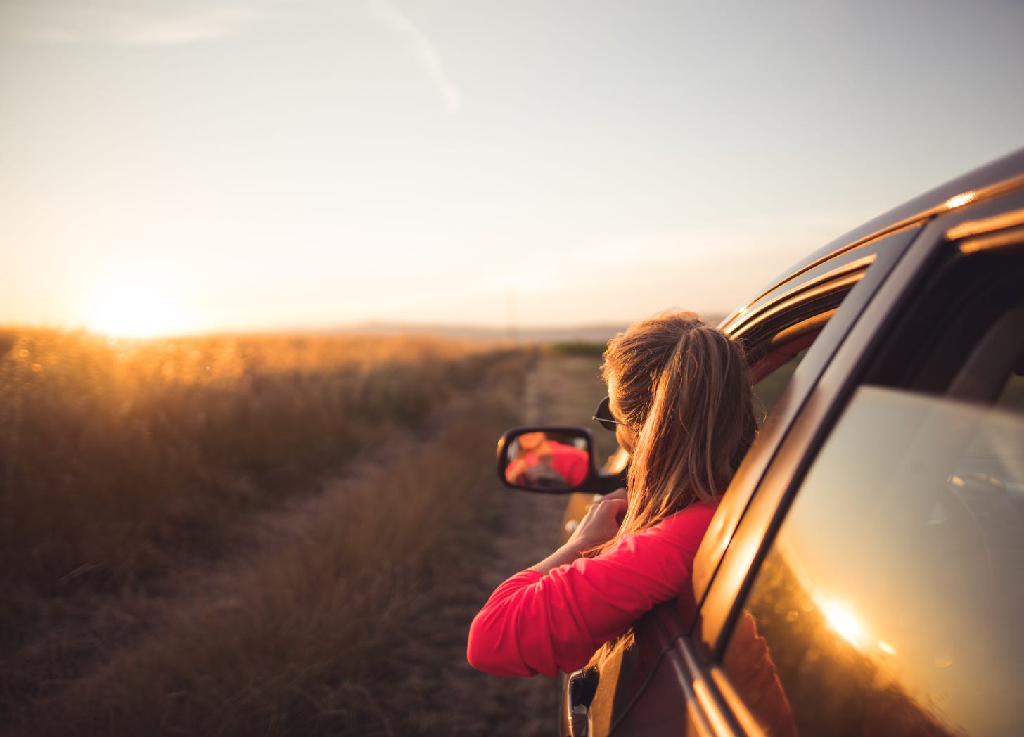 A person leaning out the window of a parked car at sunset, looking toward an open landscape and the road ahead.